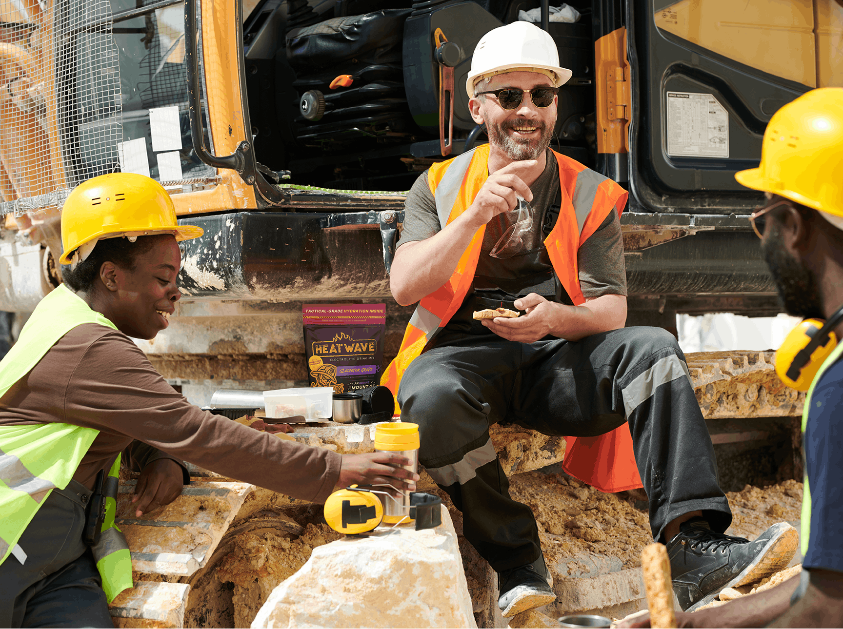 Three construction workers in hard hats and safety vests interacting near a piece of construction equipment.
